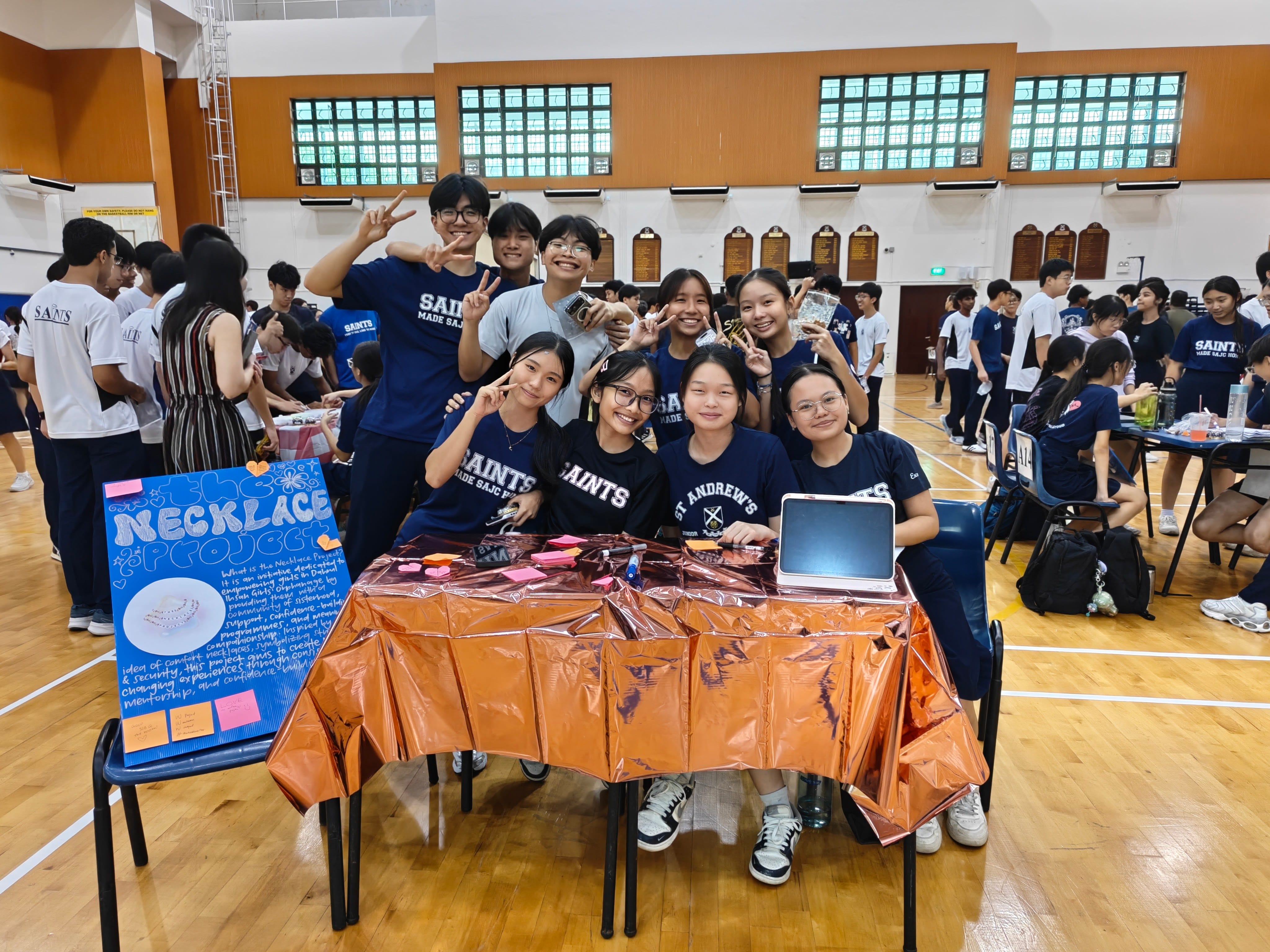A group of smiling student leaders posing for a photo behind a booth table covered in metallic foil, next to a display poster titled "The Necklace Project."